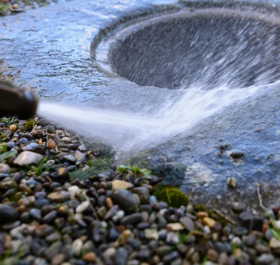 Hydro jetting process with water spraying into a sewer drain surrounded by gravel, illustrating sewer system maintenance and installation services by The Drain Fighter in Maryland.