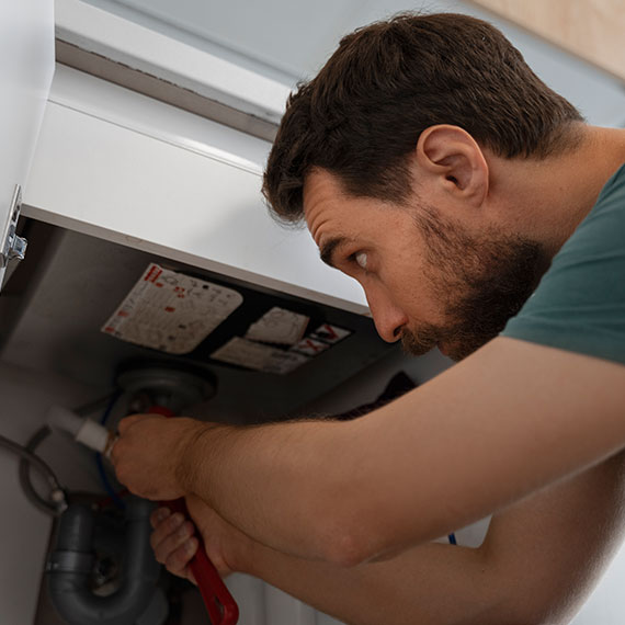 Man repairing plumbing under a sink, focusing on a water heater, illustrating urgent plumbing service needs.
